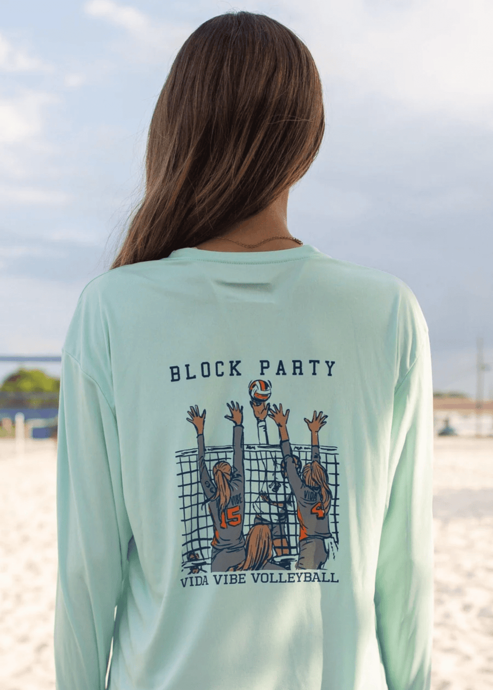 Person wearing a light green long-sleeve shirt with 'Block Party' and volleyball design on a beach.